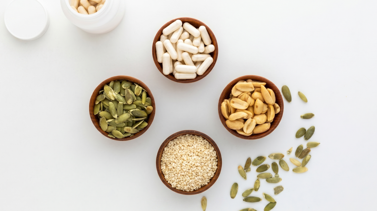 10 Sleep Aids That Actually Work: Science-Backed Options For Better Rest 10 Flat Lay View Of Four Small Wooden Bowls Arranged In A Circle On A Plain White Surface. The Bowls Contain White Glycine Capsules, Roasted Peanuts, Sesame Seeds, And Pumpkin Seeds. A White Pill Bottle With Capsules Sits In The Top Left Corner, And Loose Pumpkin Seeds Are Scattered Across The Bottom Right.