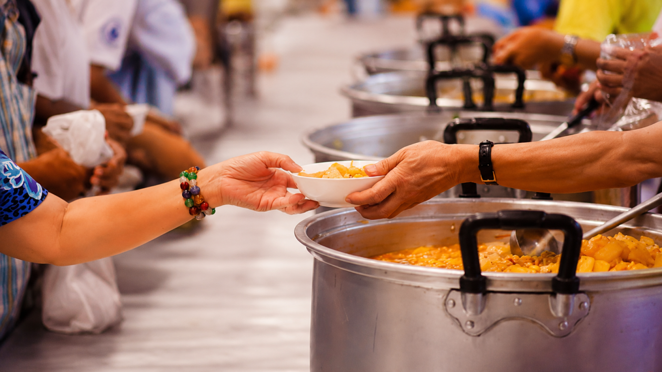What Healthy Aging Really Looks Like (And What It Doesn'T) 6 Volunteer Handing A Bowl Of Hot Food To A Person At A Community Meal Service, With Large Cooking Pots And Other Volunteers In The Background.