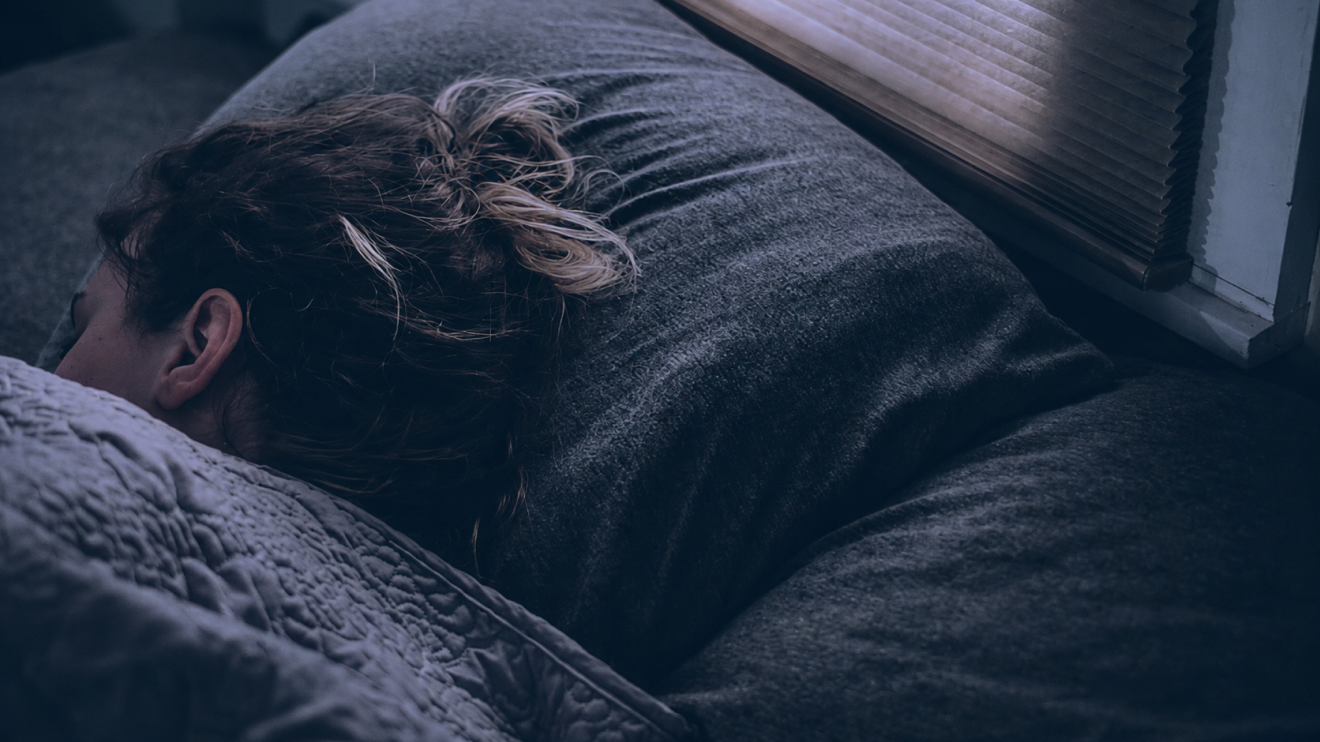 6 Healthy Habits For A Clearer, More Focused Day 2 A Woman Sleeps On Her Side Under Textured Bedding Beside A Window With Closed Blinds, In A Quiet Bedroom Scene With Cool Blue-Gray Lighting.