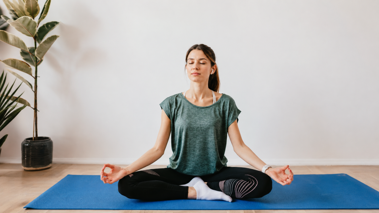 Your Simple Wellness Stack: 6 Daily Habits For Everyday Health 6 Woman Sitting Cross-Legged On A Blue Yoga Mat With Her Eyes Closed, Meditating In A Bright Minimalist Room With A Potted Plant Nearby.