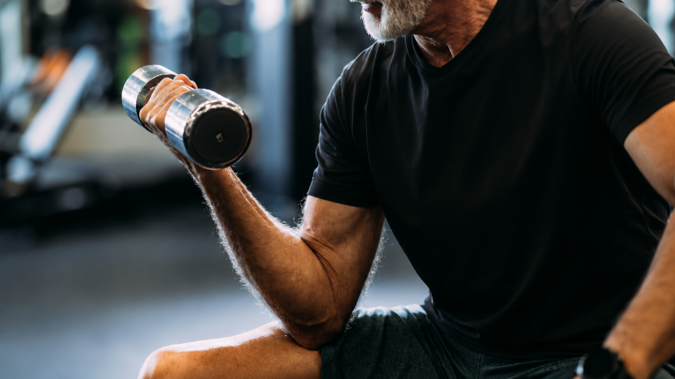 What Healthy Aging Really Looks Like (And What It Doesn'T) 2 Older Man In A Black T-Shirt Performing A Seated Dumbbell Curl In A Modern Gym.
