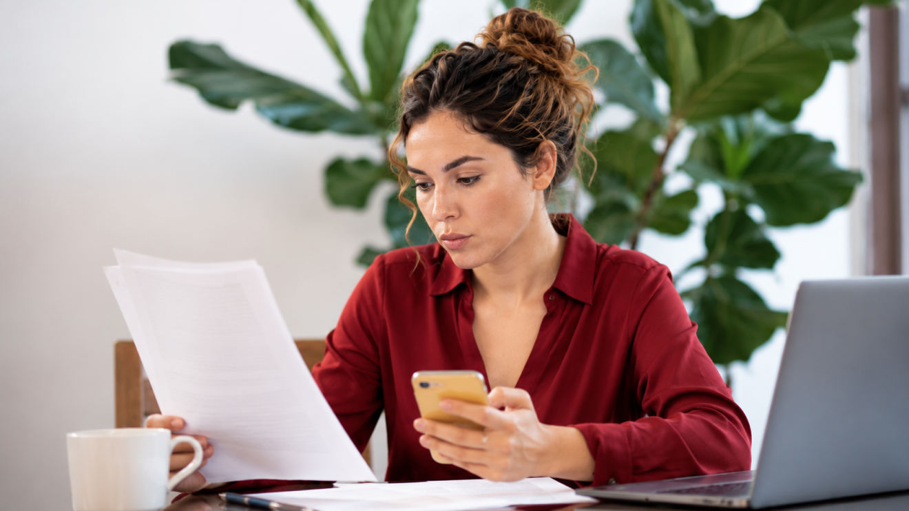 6 Healthy Habits For A Clearer, More Focused Day 6 A Woman Reviews Paperwork While Holding Her Phone Beside A Laptop, Illustrating How Digital Distractions Can Interrupt Focus During Important Work.