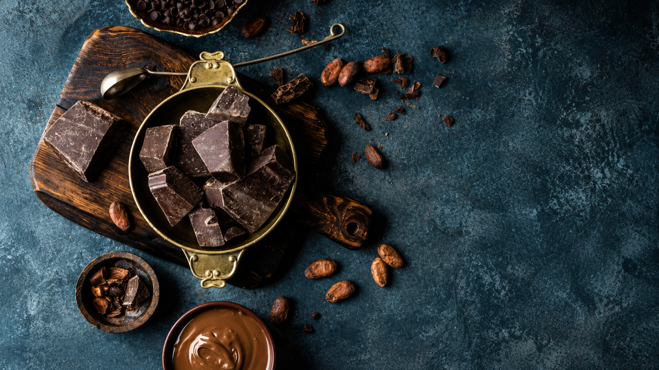 10 Foods To Eat More Often To Support Healthy Aging 5 Dark Chocolate Chunks In A Vintage Brass Bowl On A Wooden Board, Surrounded By Cacao Beans, Chocolate Chips, And Melted Chocolate On A Textured Blue Surface.