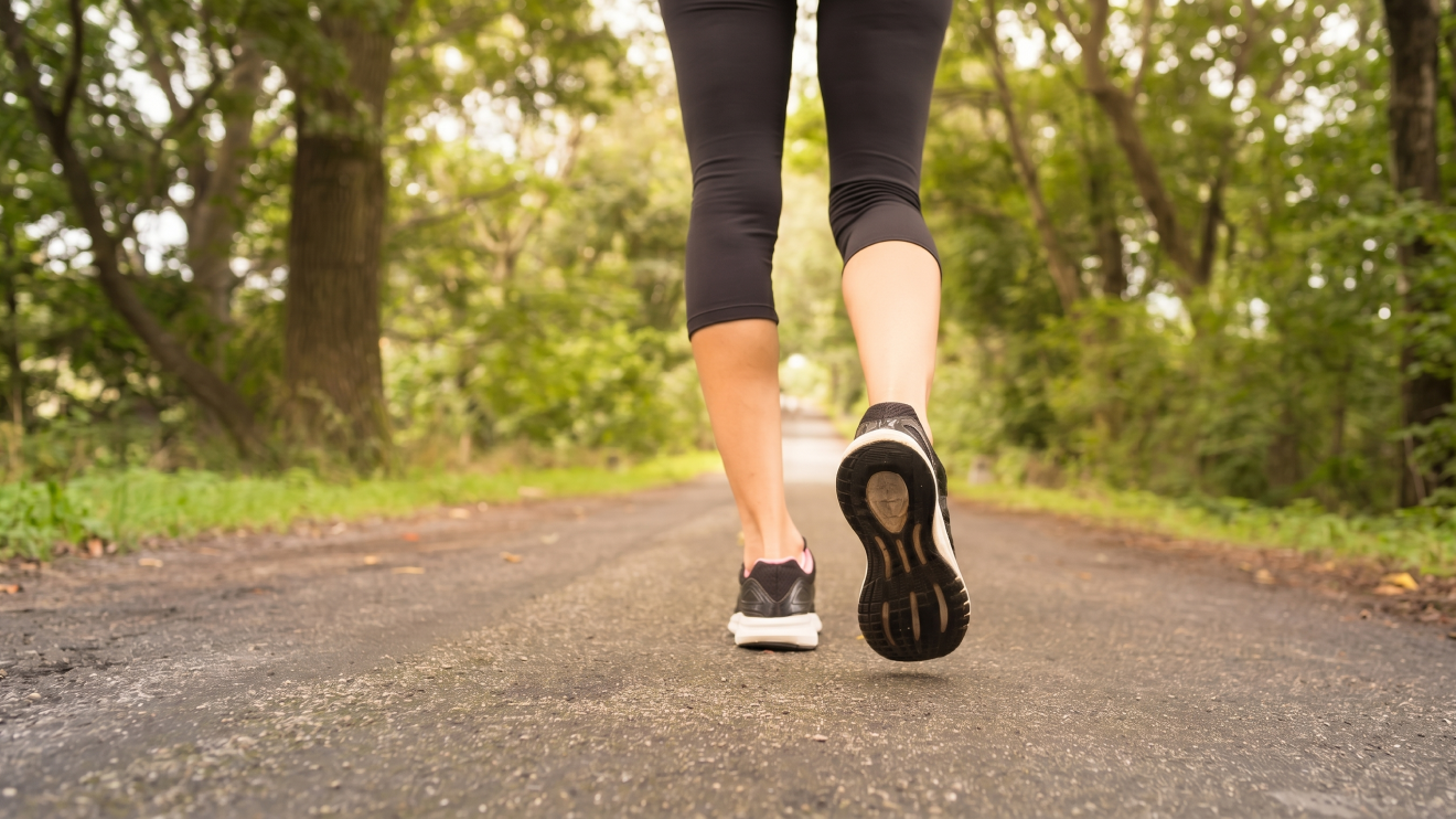 Morning Glow Routine: 8 Daily Habits For Radiant Skin 7 A Low-Angle, Rear-View Photograph Of A Person With Black Capri Leggings And Black Athletic Shoes With Pink Details, Walking On An Asphalt Path Through A Lush Green Forest. The Person Is In Mid-Stride.