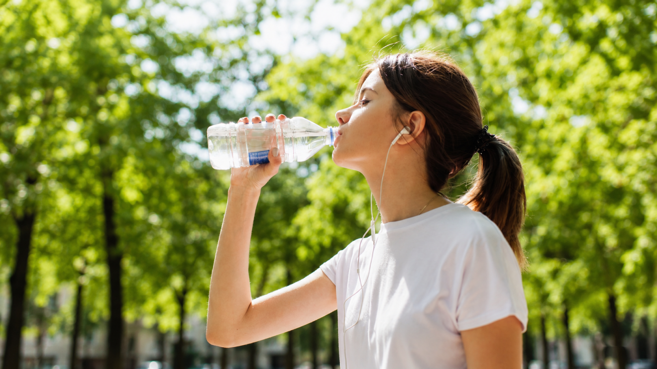 Your Simple Wellness Stack: 6 Daily Habits For Everyday Health 3 Woman In A White T-Shirt And Earbuds Drinks Water From A Plastic Bottle While Walking Outdoors In A Sunny Green Park.