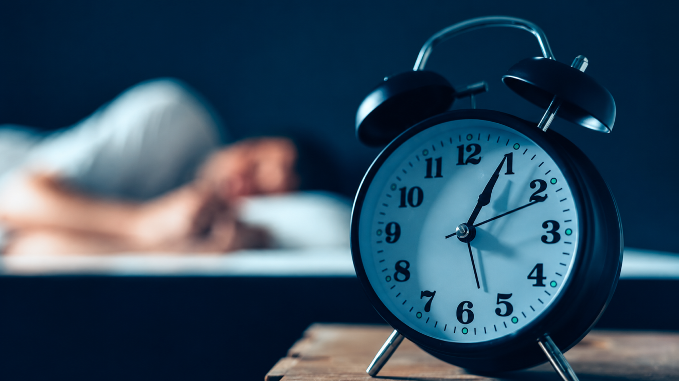 Healthy Aging Doesn'T Have To Be Complicated: 6 Simple Habits That Actually Work 5 A Black Analog Alarm Clock In Sharp Focus On A Wooden Bedside Table, With A Sleeping Person Blurred In The Background Under Cool Blue Nighttime Lighting.