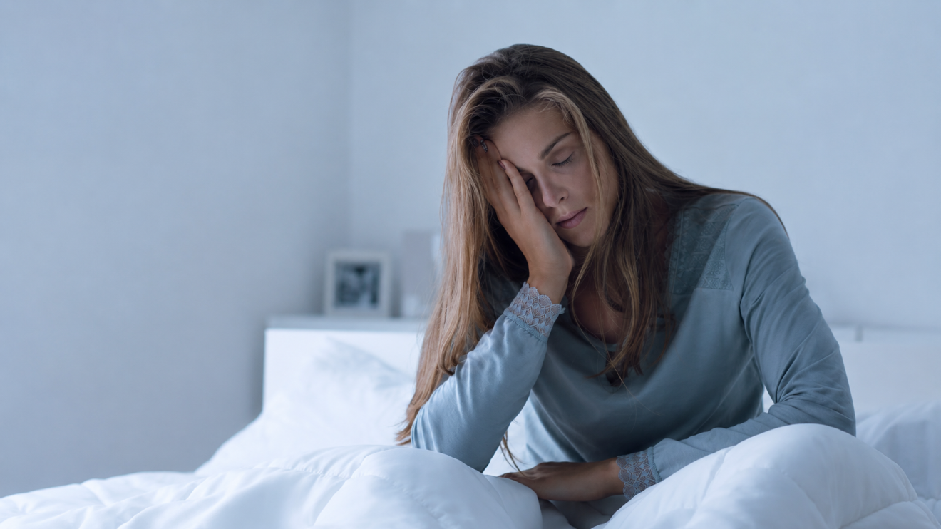 Low Energy Day Routine: 9 Steps That Still Get Results 10 Exhausted Woman Sitting In Bed In Soft Blue Morning Light, Holding Her Head While Struggling With Fatigue, Illustrating The Importance Of Prioritizing Rest Instead Of Forcing Productivity When Completely Drained. The Image Reflects How Pushing Through Exhaustion Can Harm Both Long-Term Well-Being And Future Productivity.