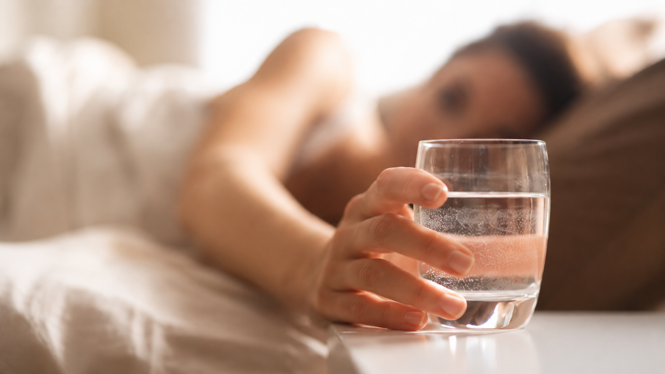 Healthy Aging Doesn'T Have To Be Complicated: 6 Simple Habits That Actually Work 2 Close-Up Of A Woman’s Hand Holding A Glass Of Water On A Bedside Table, With The Woman Resting In Bed Blurred In The Softly Lit Background.